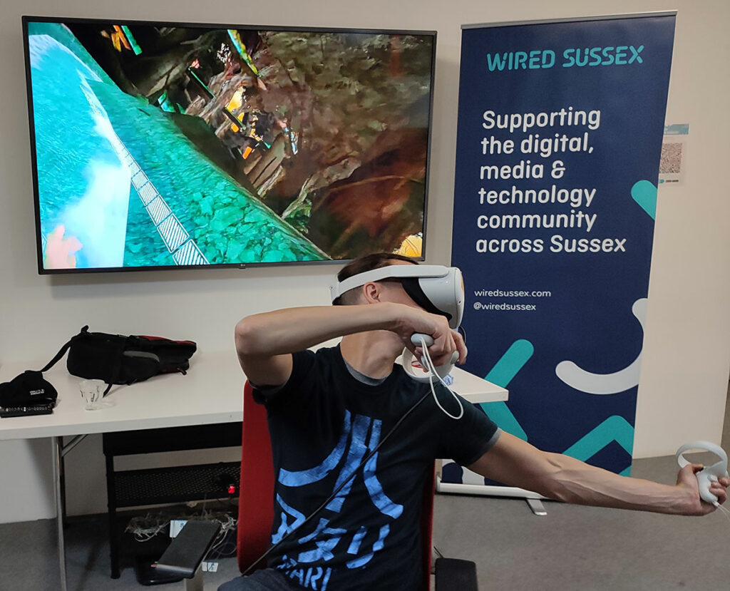 A man wearing a VR headset and holding controllers sits in a chair, sitting almost as if he is dabbing. Behind him a TV screen shows the game he is playing.