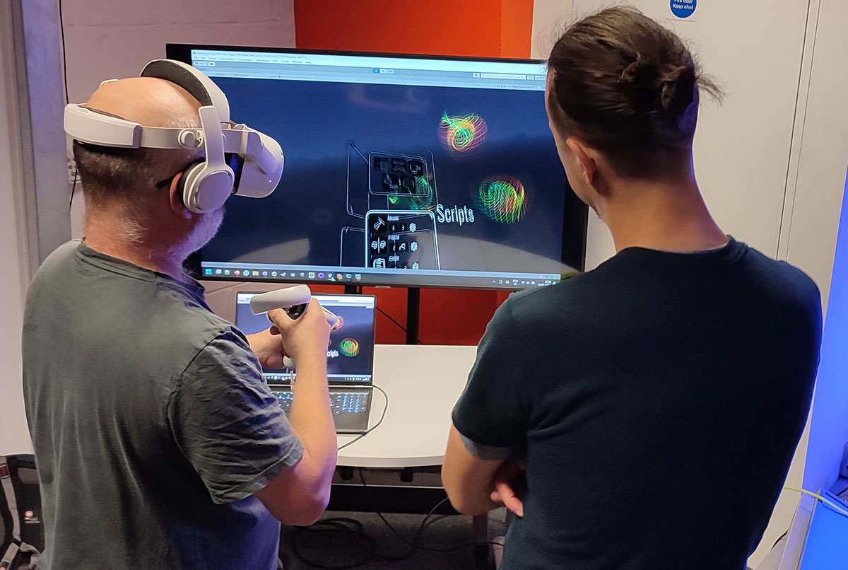 Two men stand in front of a TV screen showing a menu and multicoloured lines. One wears a VR headset and uses controllers.