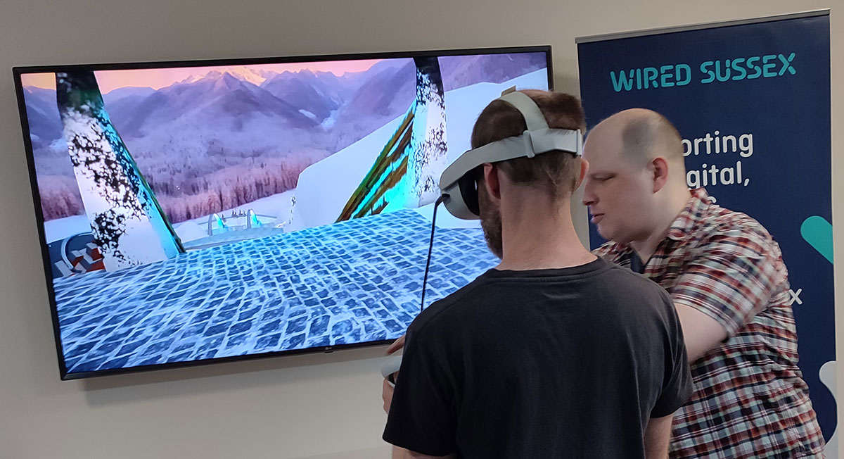A man wearing a VR headset is assisted by a man with no headset. They are standing in front of a TV showing the game played with the headset.