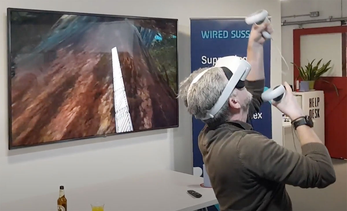 A man in a VR headset holding controllers stands looking up with his hands stretched upwards. On a TV behind him we see he is climbing a tree in a game.