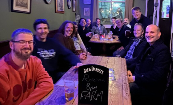 Thirteen people gathered around two tables in a pub, smiling at the camera