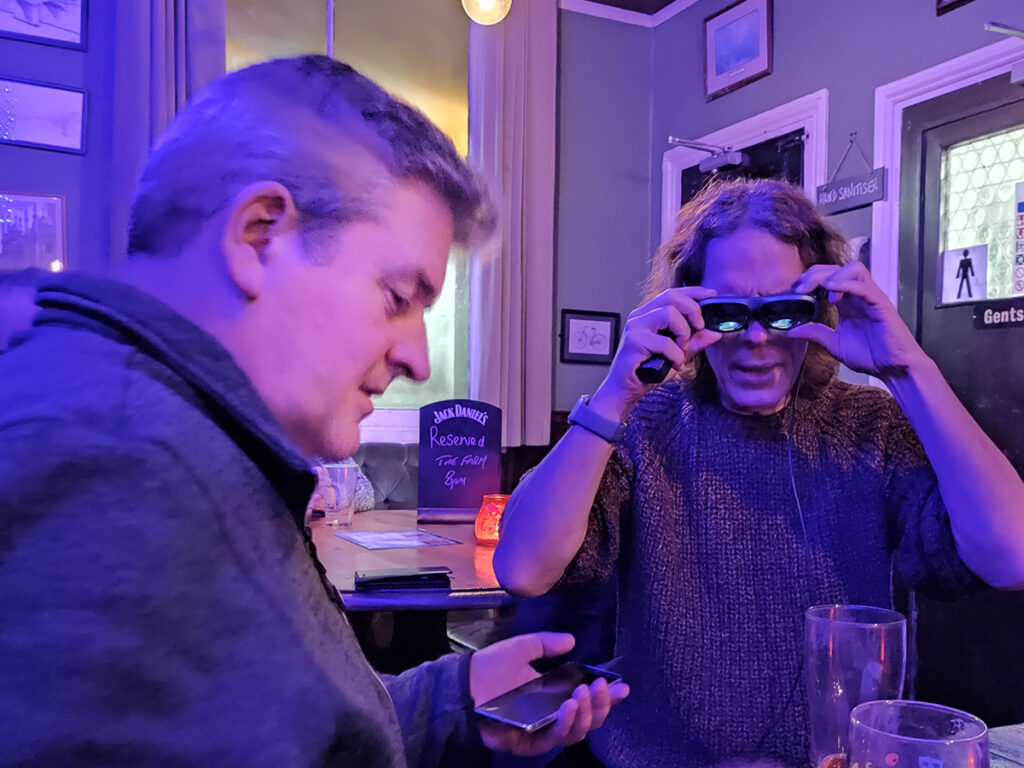 A short haired man helps a long haired man use some AR glasses attached to a phone